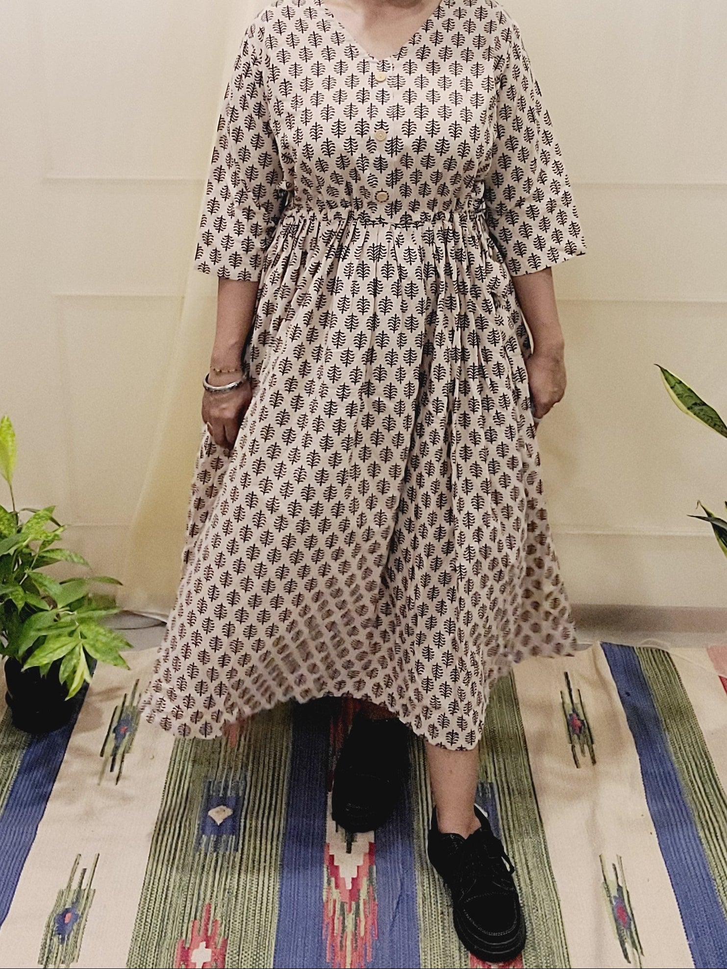 Person wearing a long flowy, beige dress with black Block printed leaves standing on a colorful rug with plants around.
