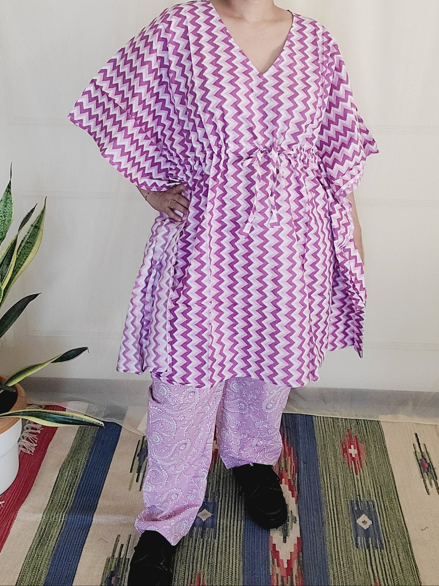 Person lying on a bed wearing a purple and white patterned dress, with a colorful rug and plant in the background.