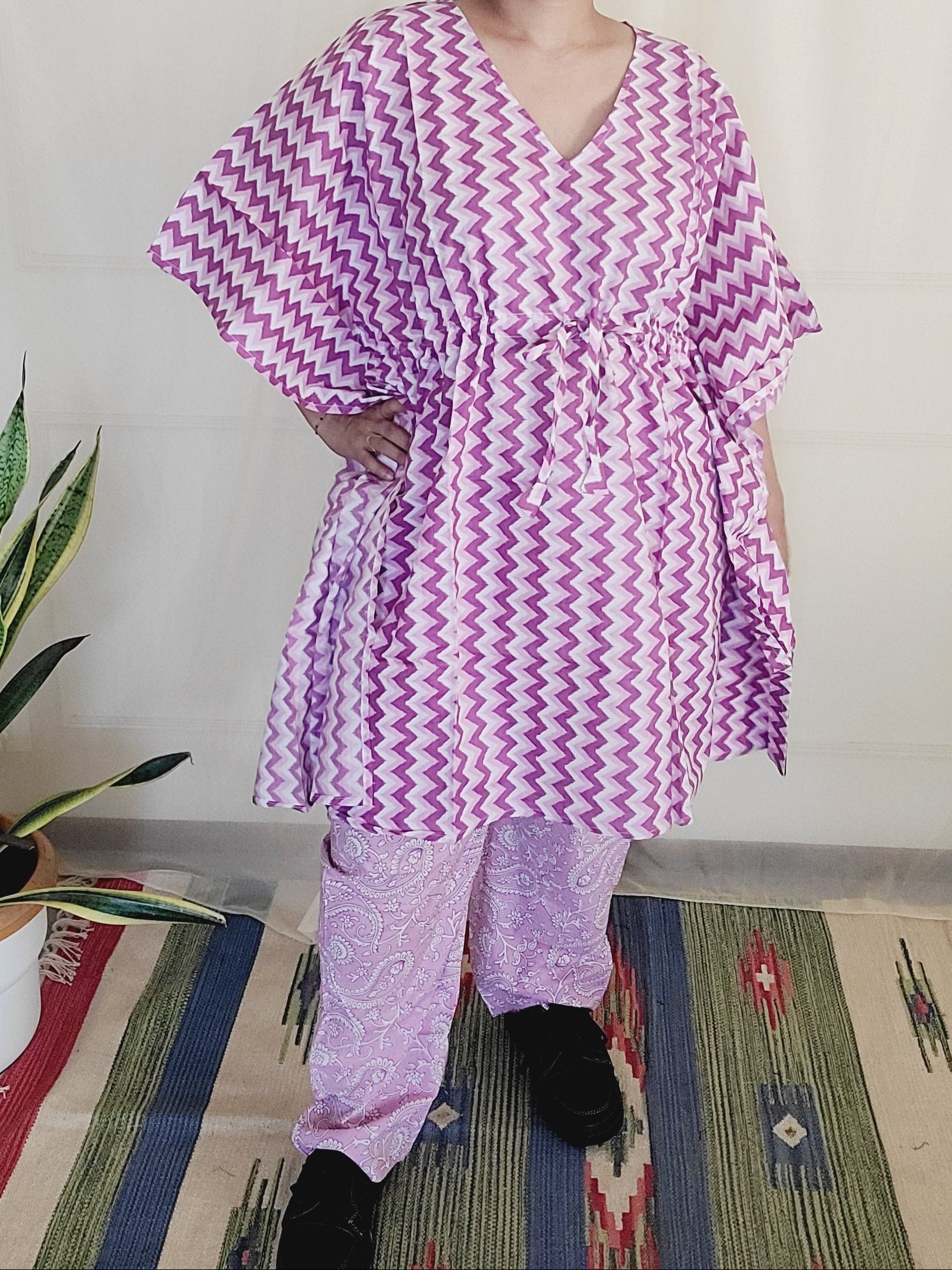 Person lying on a bed wearing a purple and white patterned dress, with a colorful rug and plant in the background.